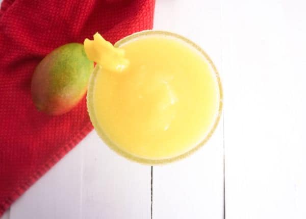 overhead view of frozen mango margarita in a glass rimmed with sugar with a piece of mango on the side with a mango on a red cloth next to it, all on a white background
