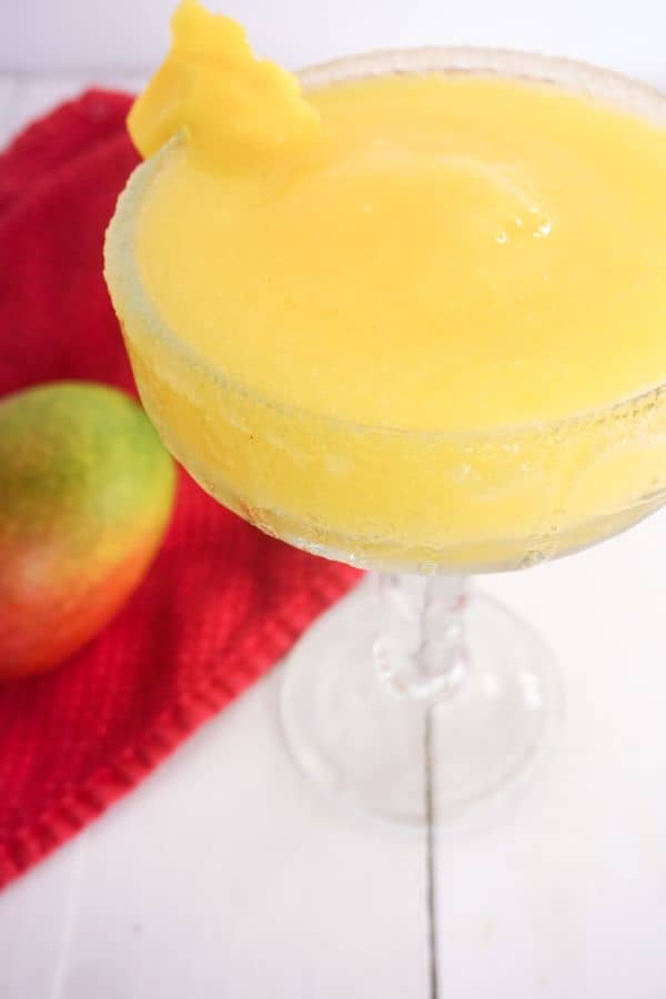 frozen mango margarita in a glass rimmed with sugar with a piece of mango on the side with a mango on a red cloth next to it, all on a white background