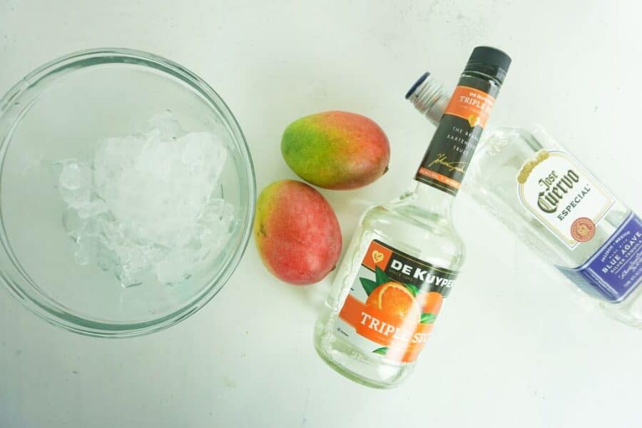 ice in a glass bowl, two mangoes, a bottle of triple sec and a bottle of tequila all on a white background