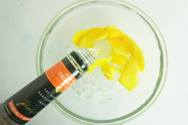 pouring triple sec into mango pieces and ice in a glass bowl on a white background