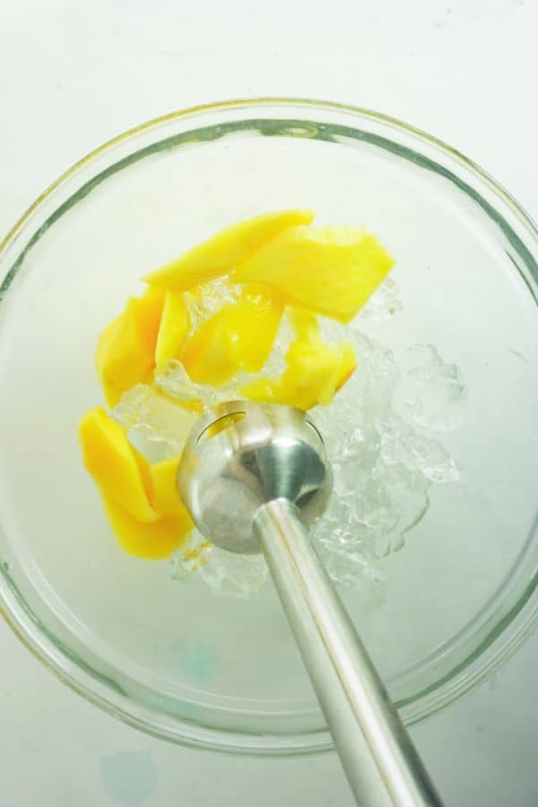 an immersion blender combining alcohol and mango pieces and ice in a glass bowl on a white background