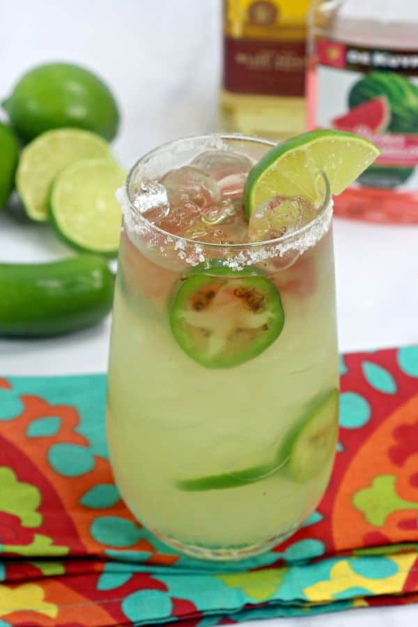 Texas Margarita with sliced jalapenos  in it and a lime wedge in a glass rimmed with salt on a turquoise and orange cloth on a white table with bottles of alcohol, jalapenos, and limes in the background