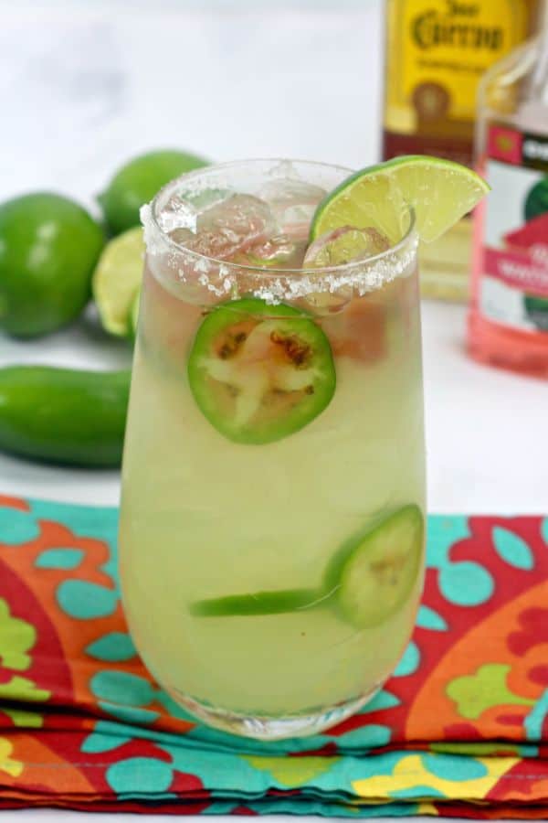 Texas Margarita with sliced jalapenos  in it and a lime wedge in a glass rimmed with salt on a turquoise and orange cloth on a white table with bottles of alcohol, jalapenos, and limes in the background