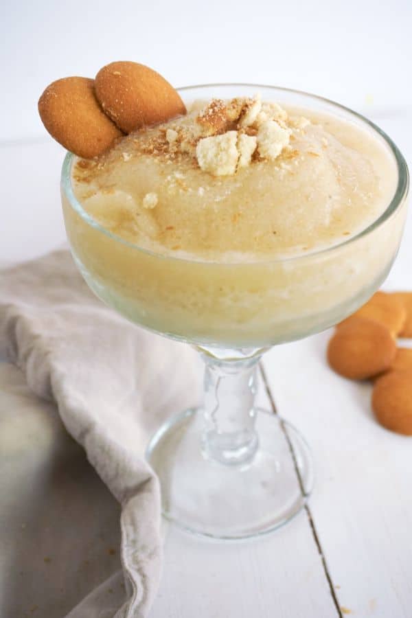 Banana Pudding Margarita in a glass next to a brown cloth and vanilla wafers on a white table