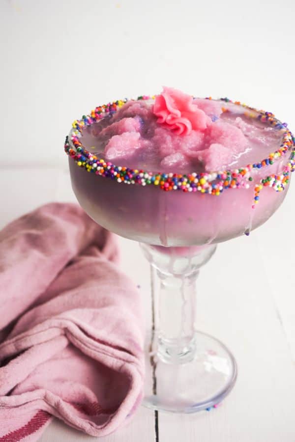 birthday cake margarita in a glass rimmed with sprinkles on a white wood table next to a pink cloth