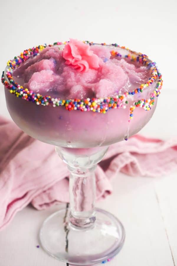 birthday cake margarita in a glass rimmed with sprinkles on a white wood table next to a pink cloth