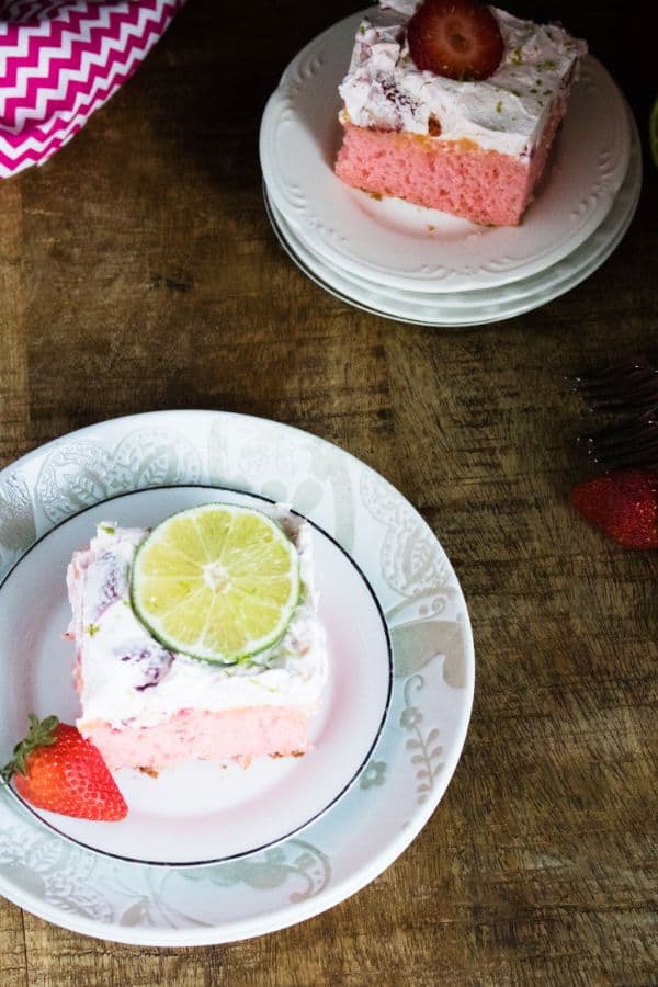 overhead view of two slices of Strawberry Margarita Poke Cake on two plates on a brown table next to a strawberry