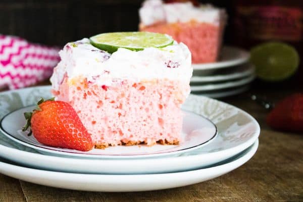 two slices of Strawberry Margarita Poke Cake on two plates on a brown table next to a strawberry, spoon, lime and bottle of alcohol 