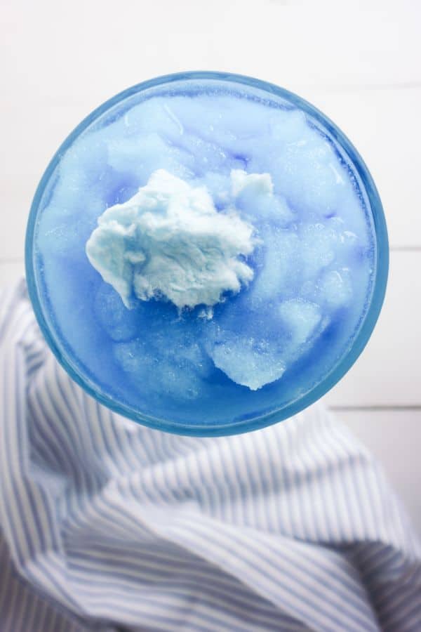 overhead view of a Cotton Candy Margarita in a glass topped with a piece of blue cotton candy on a white table next to a white and blue striped cloth
