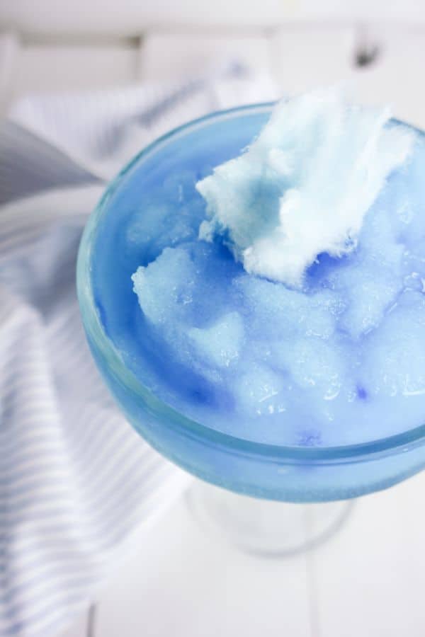 close up view of a Cotton Candy Margarita in a glass topped with a piece of blue cotton candy on a white table next to a white and blue striped cloth