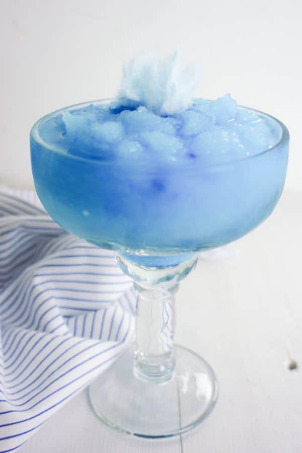 Cotton Candy Margarita in a glass topped with a piece of blue cotton candy on a white table next to a white and blue striped cloth