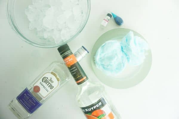 bowls of ice and blue cotton candy, bottles of tequila and triple sec, blue food coloring, and cotton candy food flavoring oil on a white background