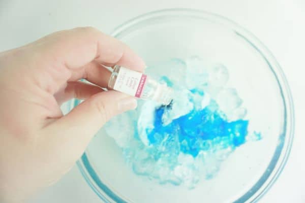 cotton candy flavoring oil being poured over blue food coloring, cotton candy and ice in a glass bowl on a white background