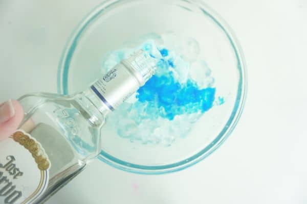 triple sec being poured over cotton candy flavoring oil, blue food coloring, cotton candy and ice in a glass bowl on a white background