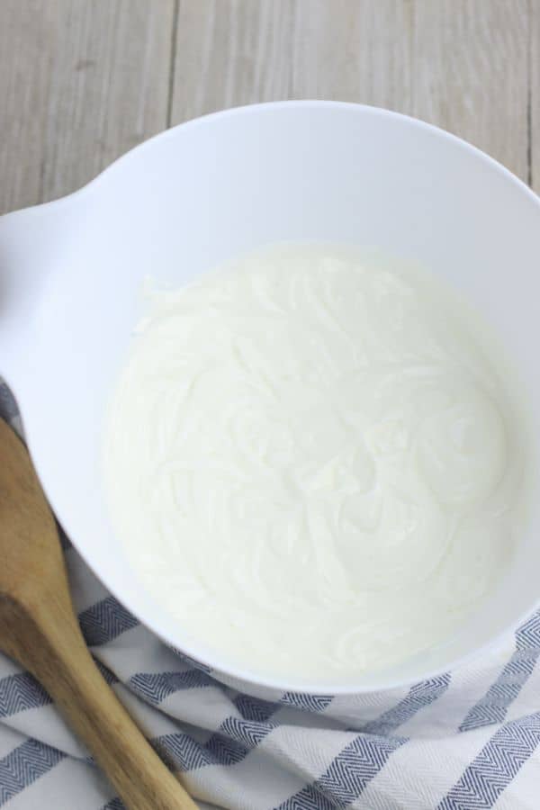 non-fat Greek yogurt in a white mixing bowl on a wood table next to a wooden spoon and a cloth