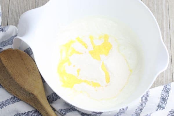 non-fat Greek yogurt, lemon pudding mix and lemon juice in a white mixing bowl on a wood table next to a wooden spoon and a cloth