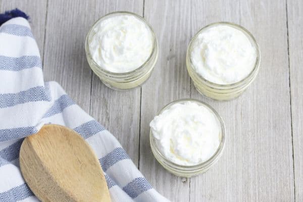 three glass jars with a vanilla wafer on the bottom of each jar topped with yogurt mixture and whipped cream on a wood table next to a wooden spoon and a cloth