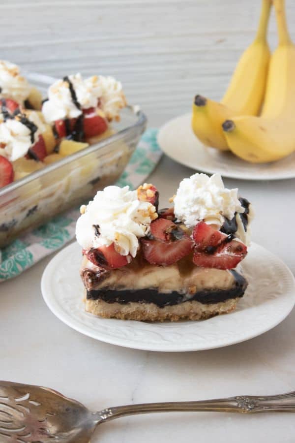 a slice of banana split cake on a white plate on a white table with more cake in a glass baking dish and bananas on a white plate in the background
