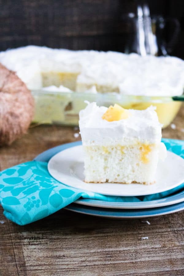 a slice of Boozy Pina Colada Cake on a plate on a brown table with more cake in a glass baking dish and a coconut in the background