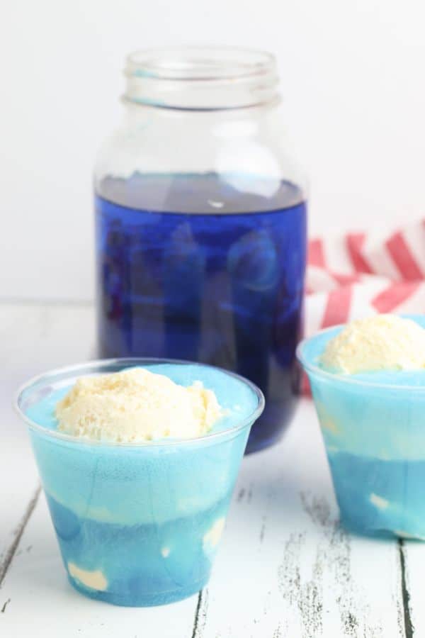 two glasses of Ocean Water Ice Cream Float on a white table next to a glass jar of lime juice colored blue and a red and white striped cloth