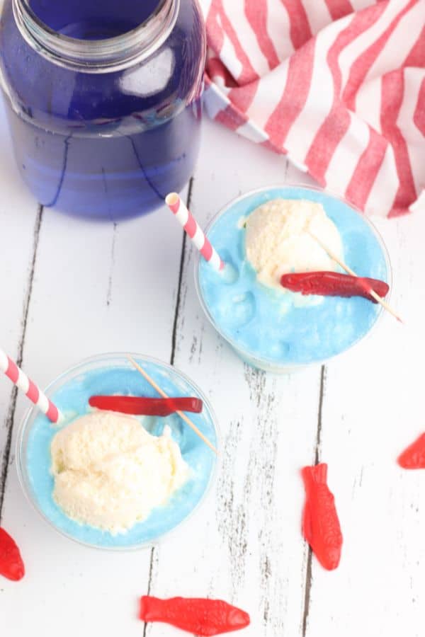 overhead view of two glasses of Ocean Water Ice Cream Float on a white table next to a glass jar of lime juice colored blue and a red and white striped cloth