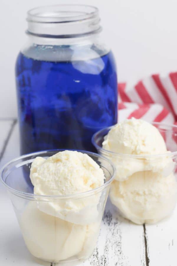 a glass jar filled with lime soda colored blue next to two cups of vanilla icing next to a red and white striped cloth on a white wood table