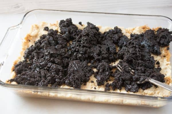 graham cracker crumbs in a glass baking dish topped with a crumble mixture and oreo cookie mixture on a white counter