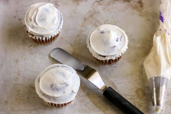 cupcakes with vanilla frosting on them next to a spreading spatula and pastry bag with icing in it
