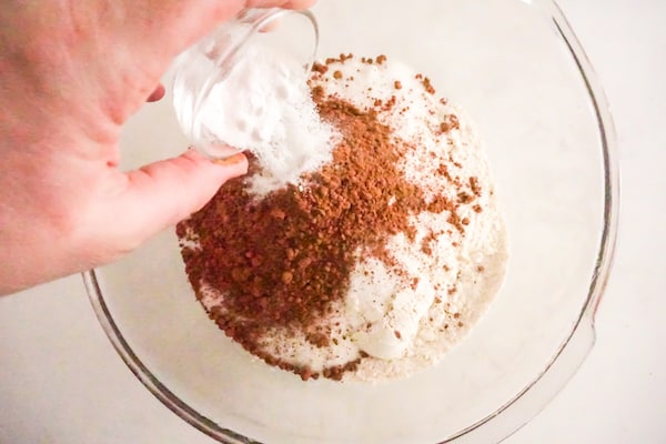 a hand pouring dry ingredients for cookies and cream cupcakes into a glass mixing bowl